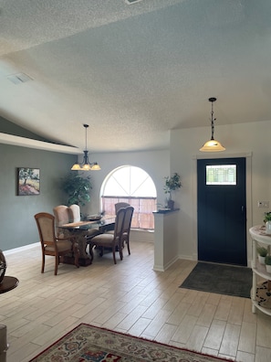 A dining area with a table, chairs, and pendant lights. There is a large window with a view, a door, and a potted plant.