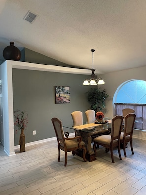 A dining area with a glass table, wooden chairs, a chandelier, and a large window.