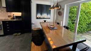 A modern kitchen with dark cabinets, a wooden dining table, pendant lights, and a view of greenery outside.