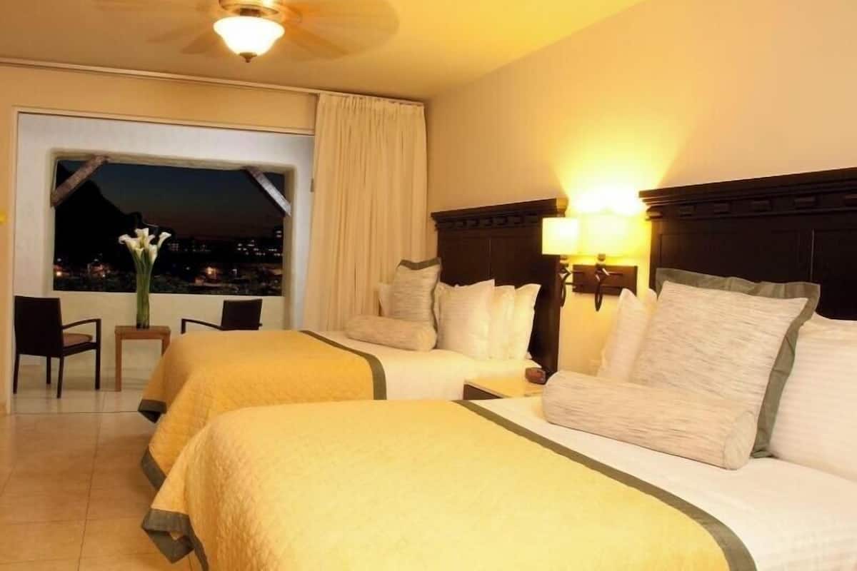 Une chambre d’hôtel avec deux lits, un ventilateur de plafond, une vue sur la ville la nuit depuis la fenêtre et une chaise près de la fenêtre.