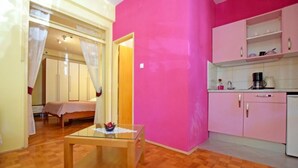 A room with a bed, a glass-top table with a bowl of fruit, a pink wall, and a kitchen area with pink cabinets and a white countertop. - Apartments Adriana - Studio Apartment A2+1 (Starigrad-Paklenica)