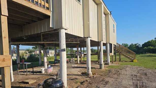 Edificio en construcción con vigas y columnas de madera a la vista, una parrilla y suelo de tierra.