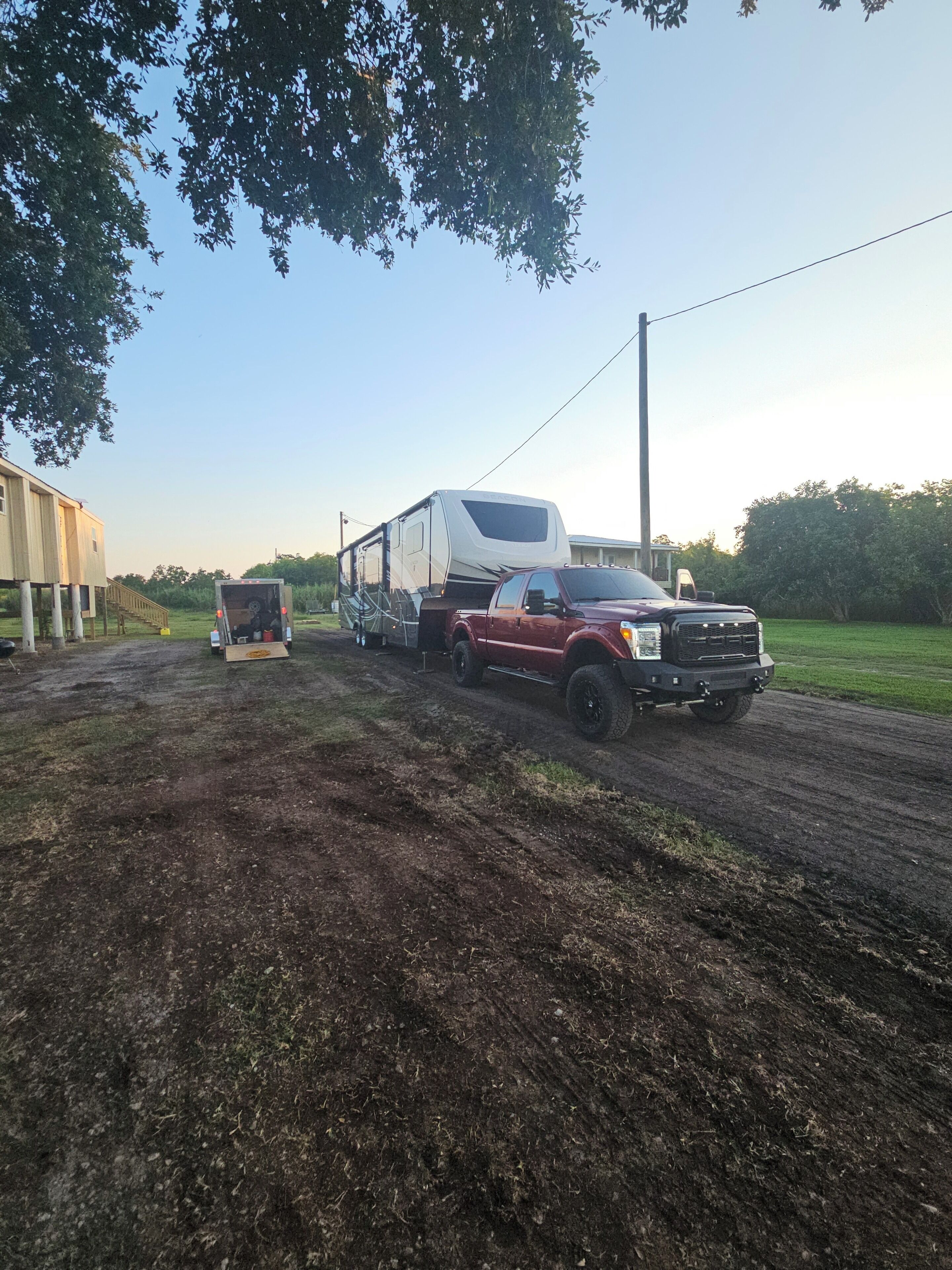 A red truck towing a white RV on a dirt road with a house in the background.
