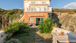 A two-story building with a balcony, a green door, and a garden area with plants and a table. - Magic Seaside Apartment (Rosignano Marittimo)