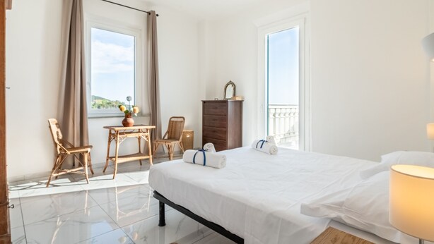 A hotel room with a bed, a desk with chairs, a dresser, and a view of the outdoors. - Magic Seaside Apartment (Rosignano Marittimo)