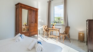 A hotel room with a large bed, a wooden wardrobe, a small table with a vase, two wicker chairs, and a window with a view of greenery. - Magic Seaside Apartment (Rosignano Marittimo)