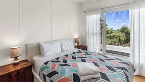 A bedroom with a bed, a nightstand, a lamp, and a view of greenery through a window. - Ocean View Skenes Creek Beach House (Skenes Creek)