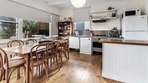 A kitchen with a dining table, chairs, a refrigerator, a stove, and a hanging light fixture. - Ocean View Skenes Creek Beach House (Skenes Creek)