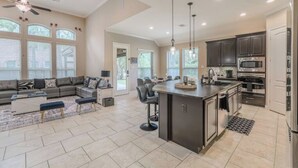A spacious living area with a large sofa, a coffee table, and a dining area with a bar. There is a kitchen with dark cabinets and stainless steel appliances.