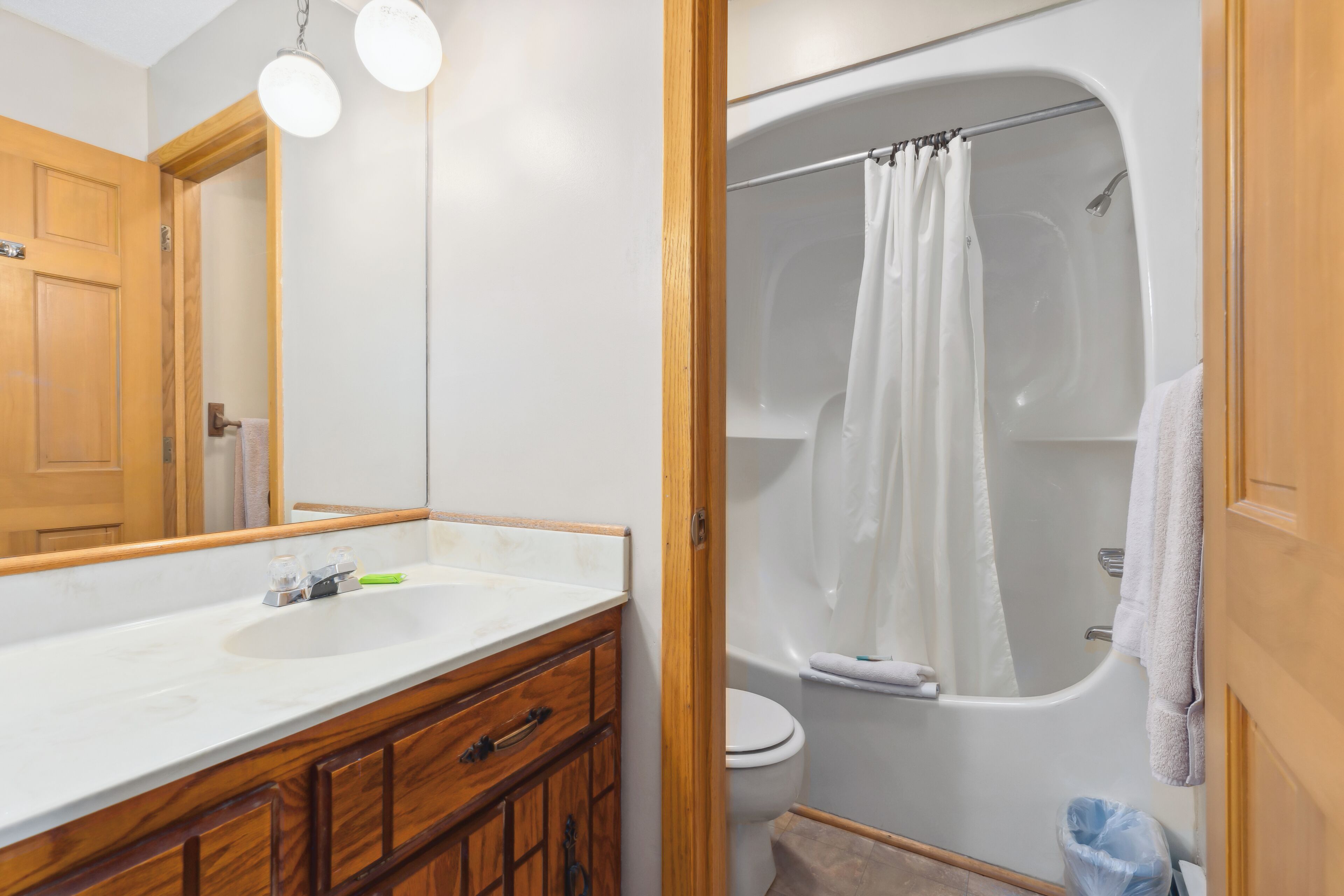 Un baño con lavamanos blanco, tocador de madera, un espejo, ducha con cortina y un inodoro.