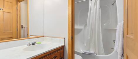 Un baño con lavamanos blanco, tocador de madera, un espejo, ducha con cortina y un inodoro.