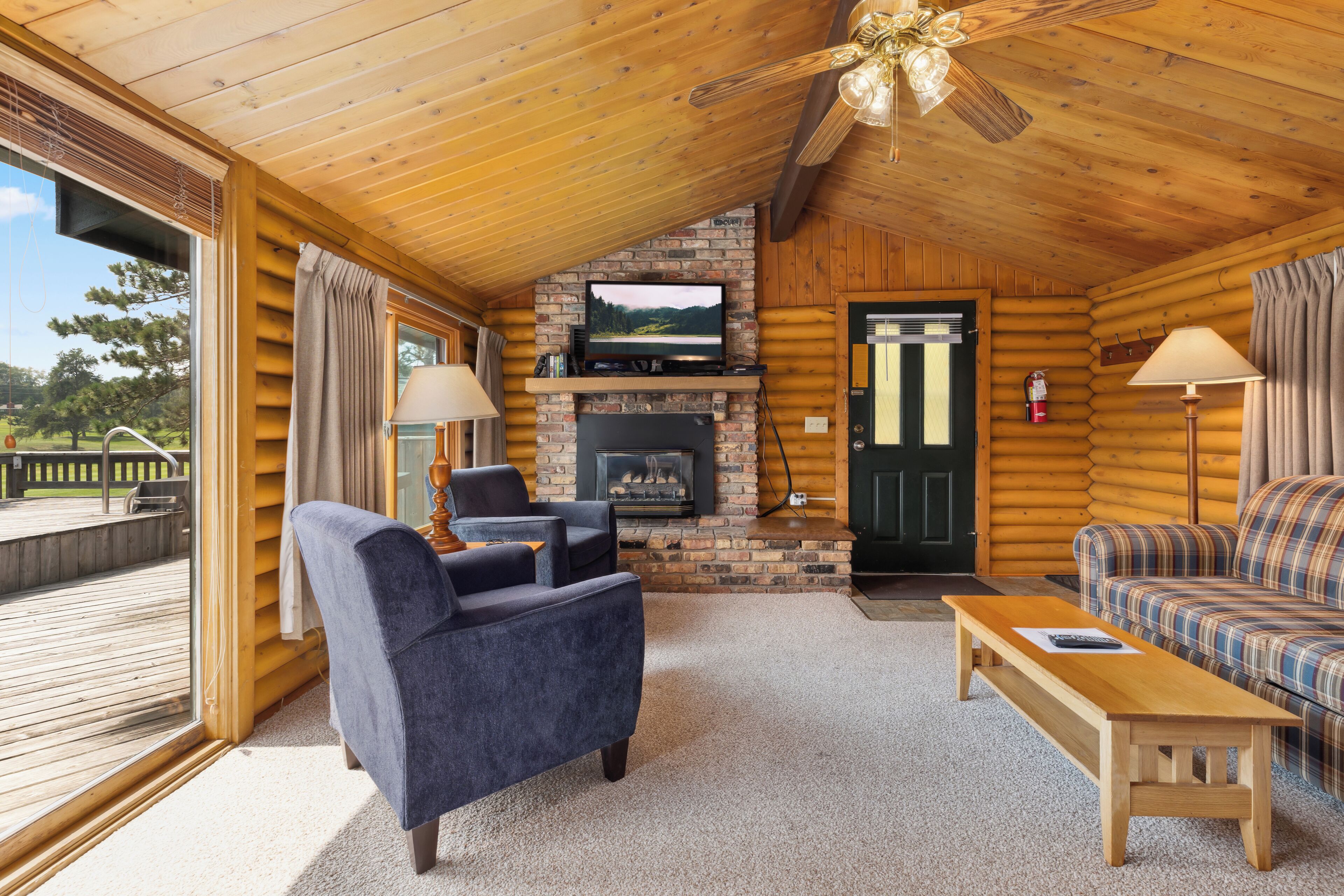 A log cabin interior with a stone fireplace, a television mounted above it, a wooden ceiling fan, and a view of a deck through a large window.