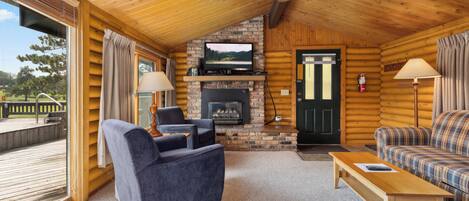 A log cabin interior with a stone fireplace, a television mounted above it, a wooden ceiling fan, and a view of a deck through a large window.