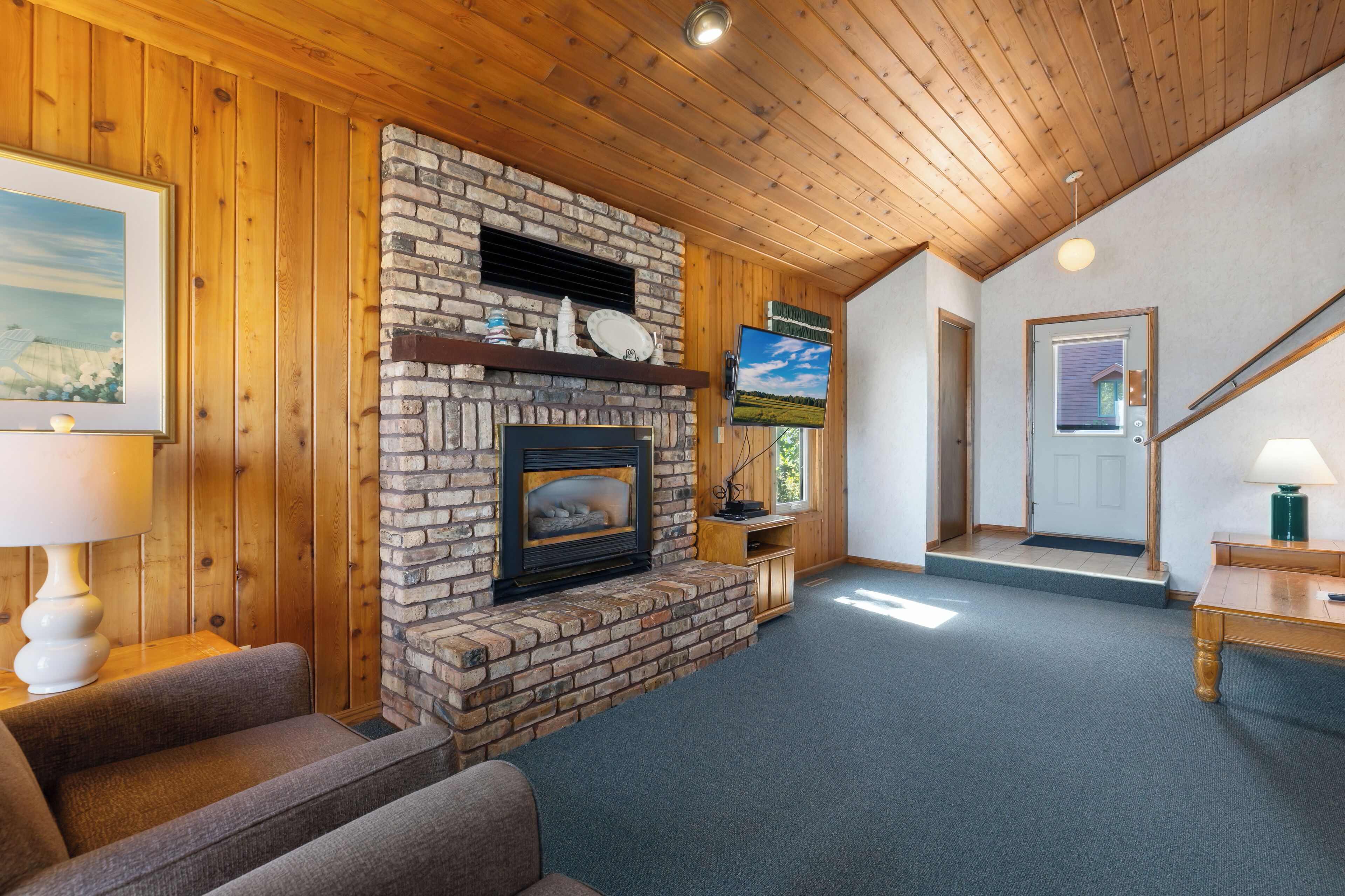A cozy living room with a brick fireplace, wooden walls, a comfortable sofa, and a television mounted on the wall.