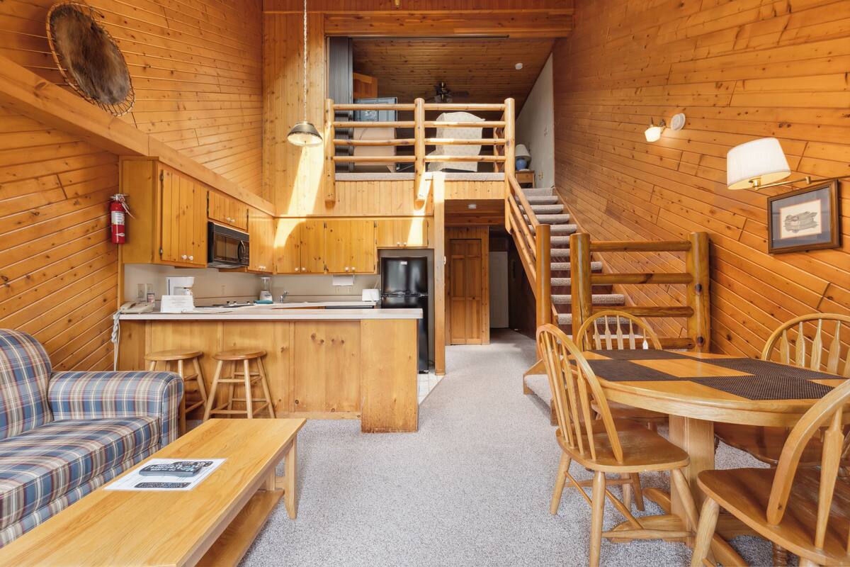 A cozy cabin interior with a kitchen, dining area, and a loft bedroom.