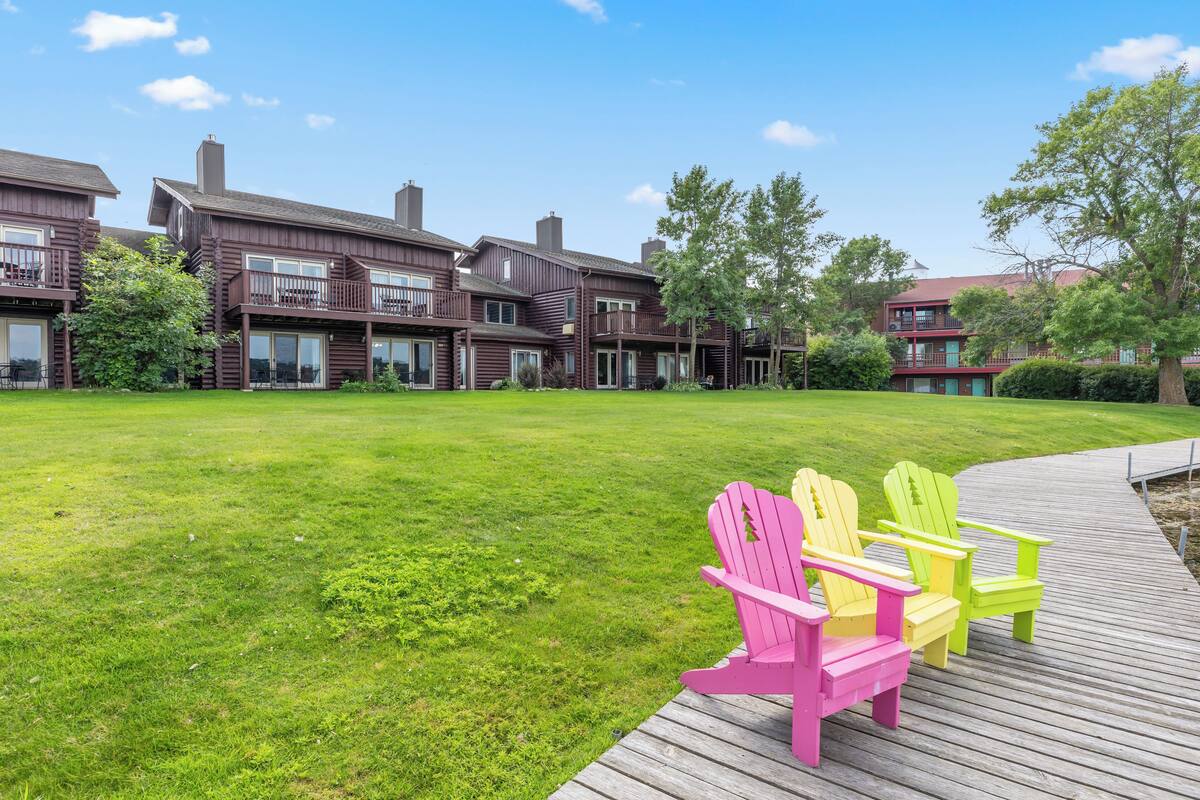 A residential area with wooden houses, a grassy lawn, and a wooden deck with colorful Adirondack chairs.