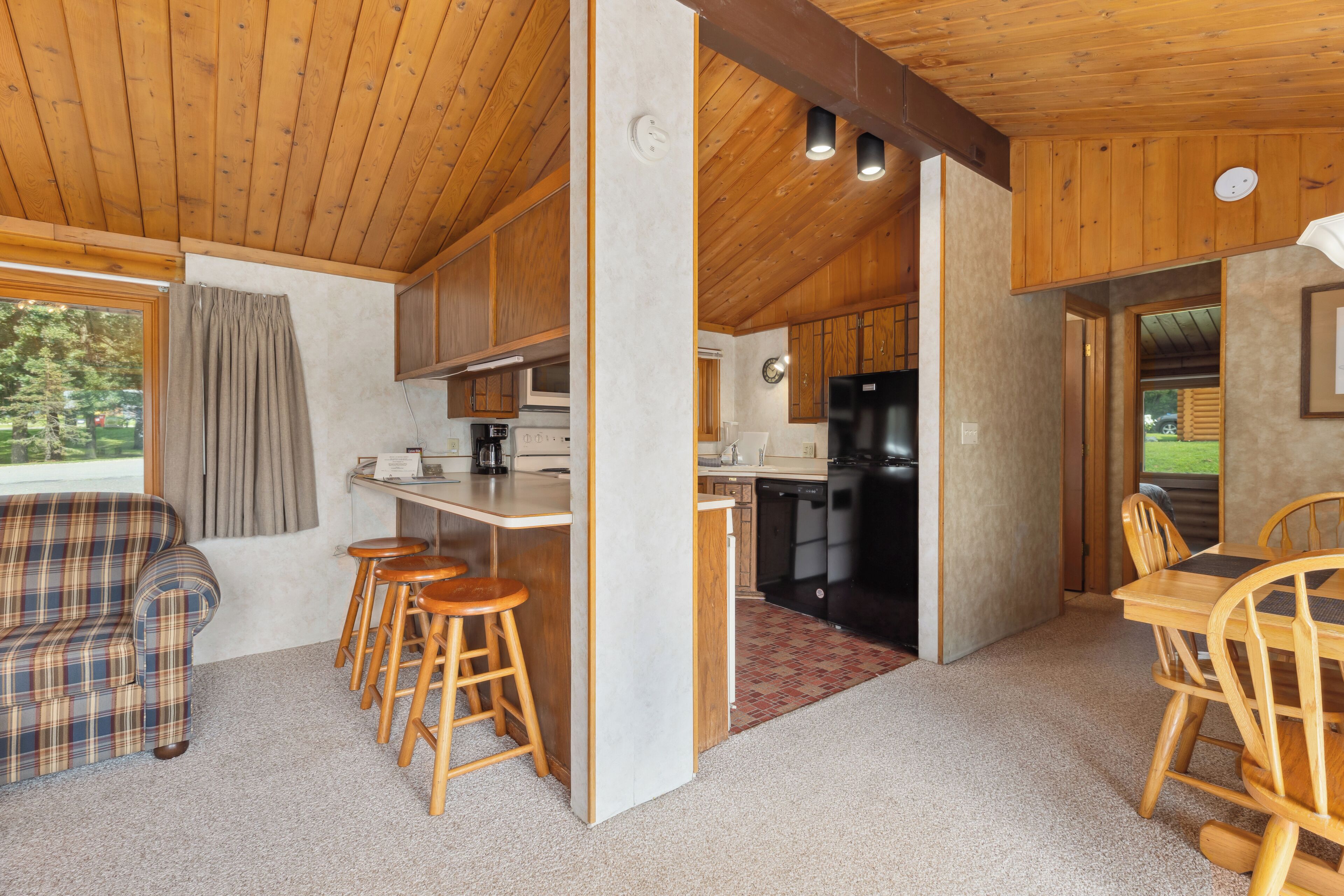 A cozy cabin interior with a wooden ceiling, a kitchenette, a dining area with wooden chairs, and a plaid sofa.