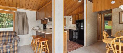 A cozy cabin interior with a wooden ceiling, a kitchenette, a dining area with wooden chairs, and a plaid sofa.