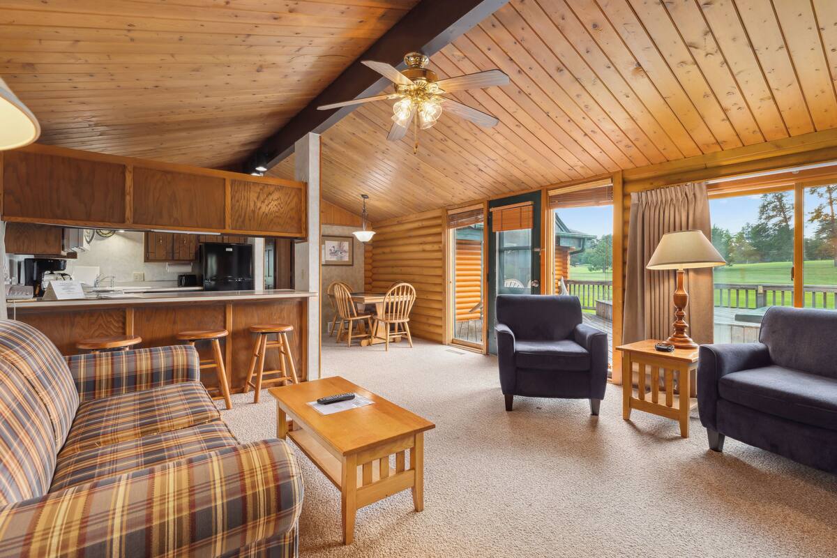 A cozy cabin interior with a wooden ceiling, a dining area, a living room with a sofa and chairs, and large windows offering a view of the outdoors.