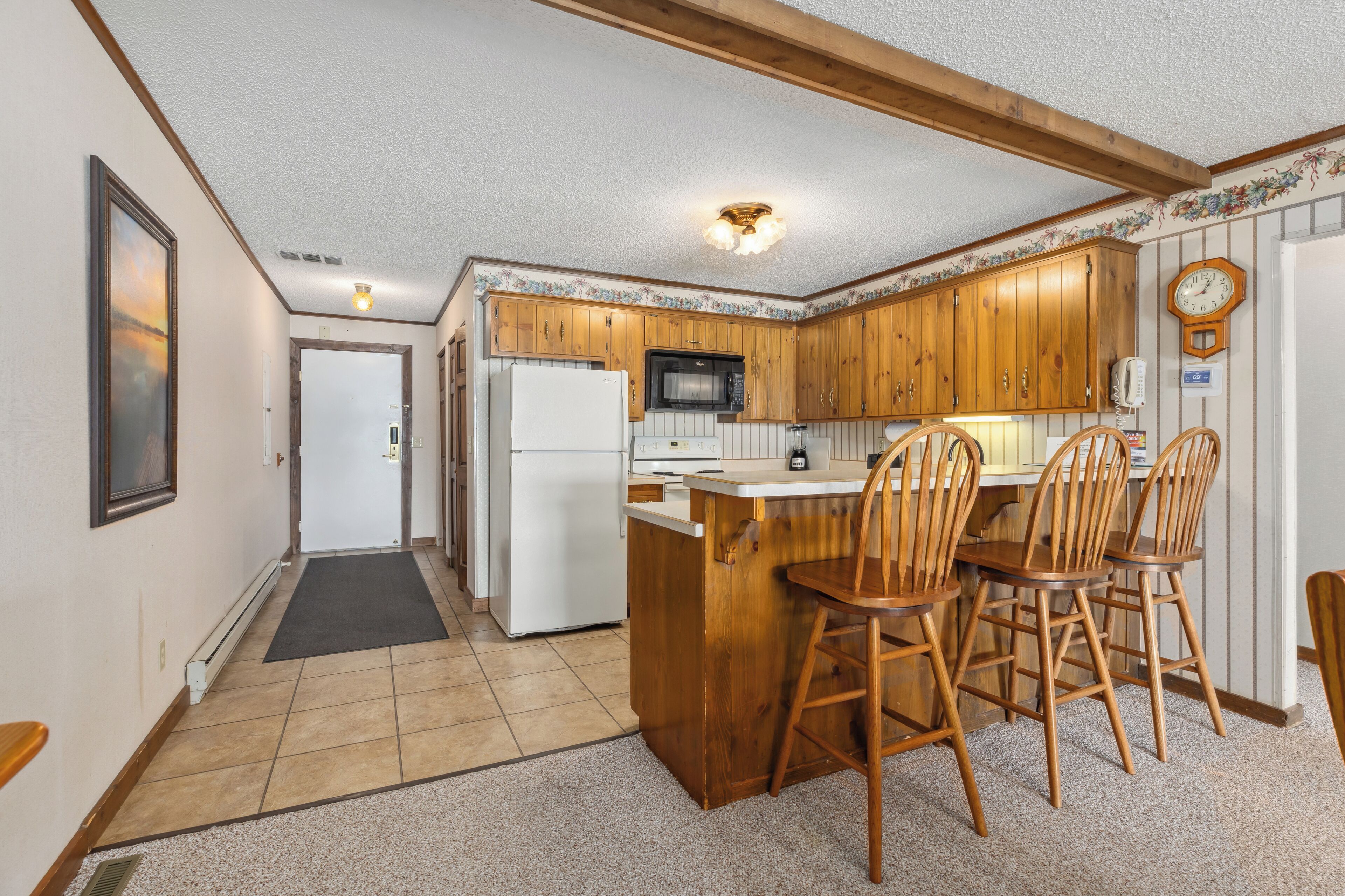 A kitchen with wooden cabinets, a refrigerator, a microwave, and a sink with wooden chairs.