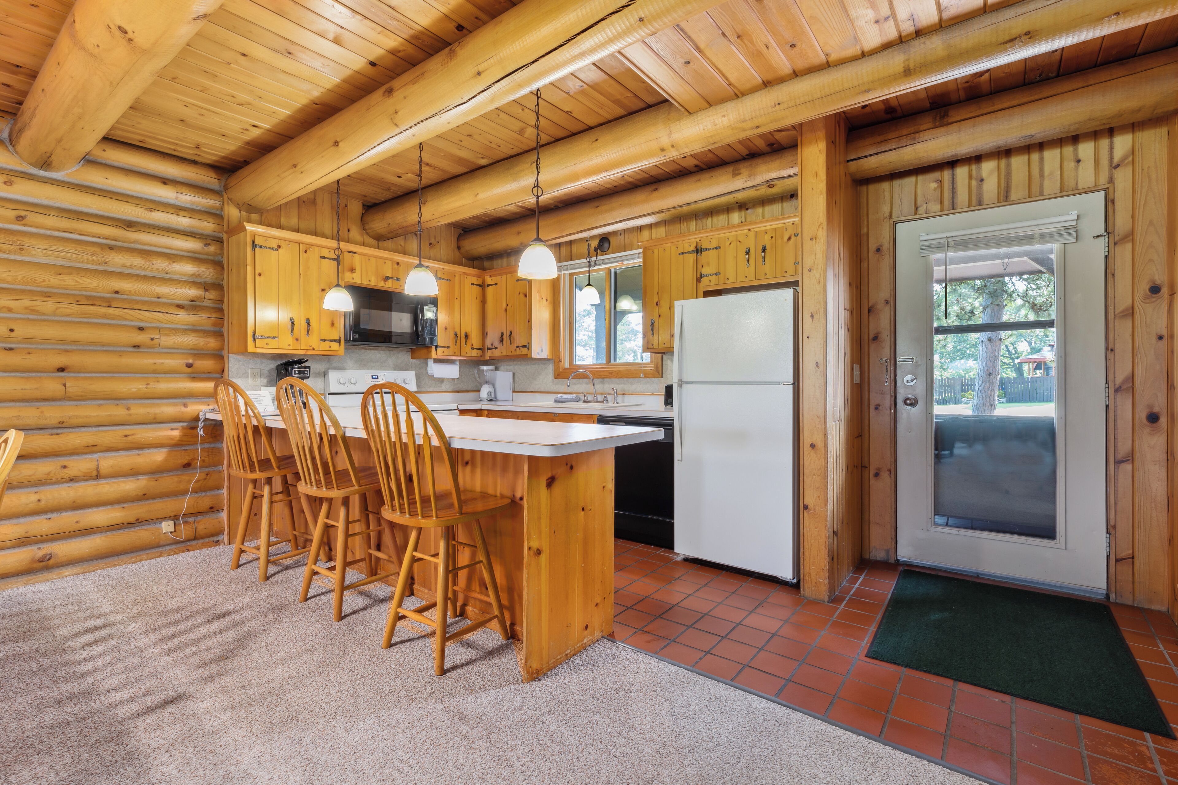 A wooden cabin kitchen with a central island, wooden chairs, a refrigerator, and a door leading outside.