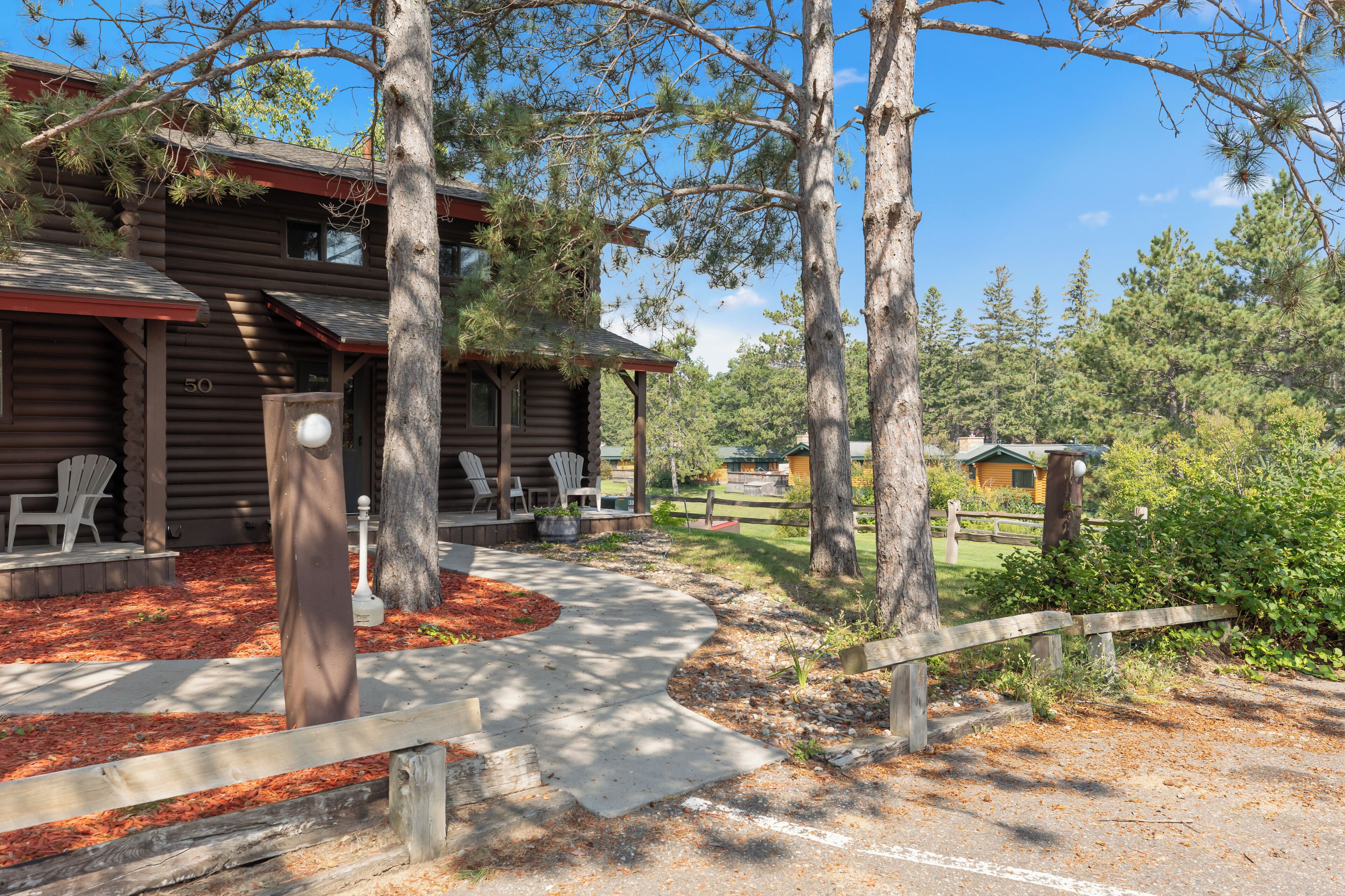 Un chalet rustique en rondins de bois, doté d’une véranda, d’un espace extérieur aménagé pour s’asseoir et d’un jardin bien entretenu.