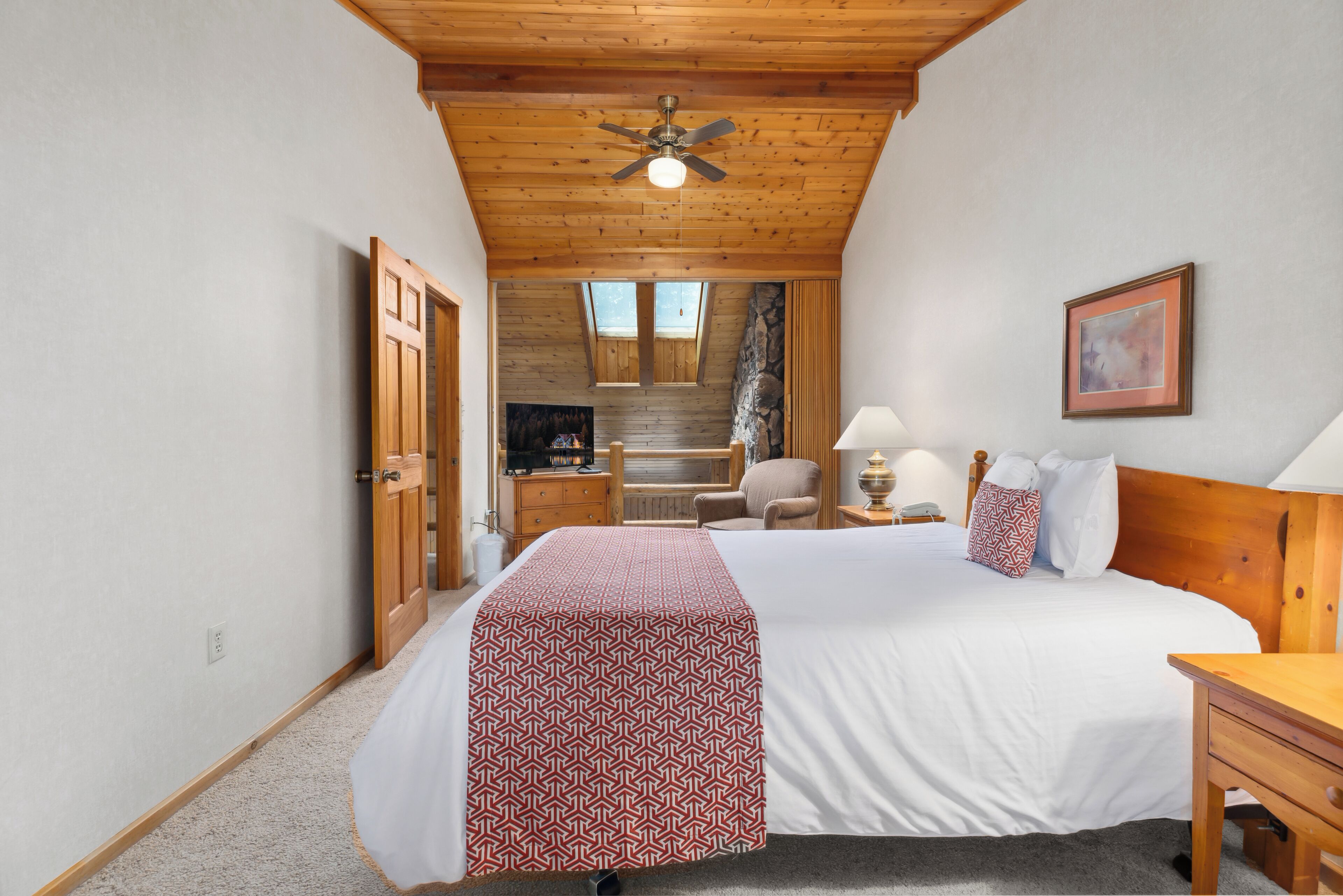 Een slaapkamer met een houten plafond, een bed met een geborduurde sprei, een nachtkastje, een stoel en een schilderij aan de muur.