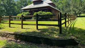 Una valla de fusta rústica envolta una casa amb façana vermella i teulat inclinat, situada en un paisatge verd i abundant.