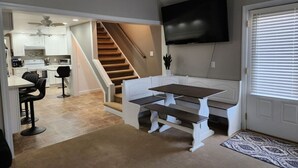 Een ruime woonkamer met een trap naar een keuken, een eettafel met banken en een flatscreen televisie aan de muur.