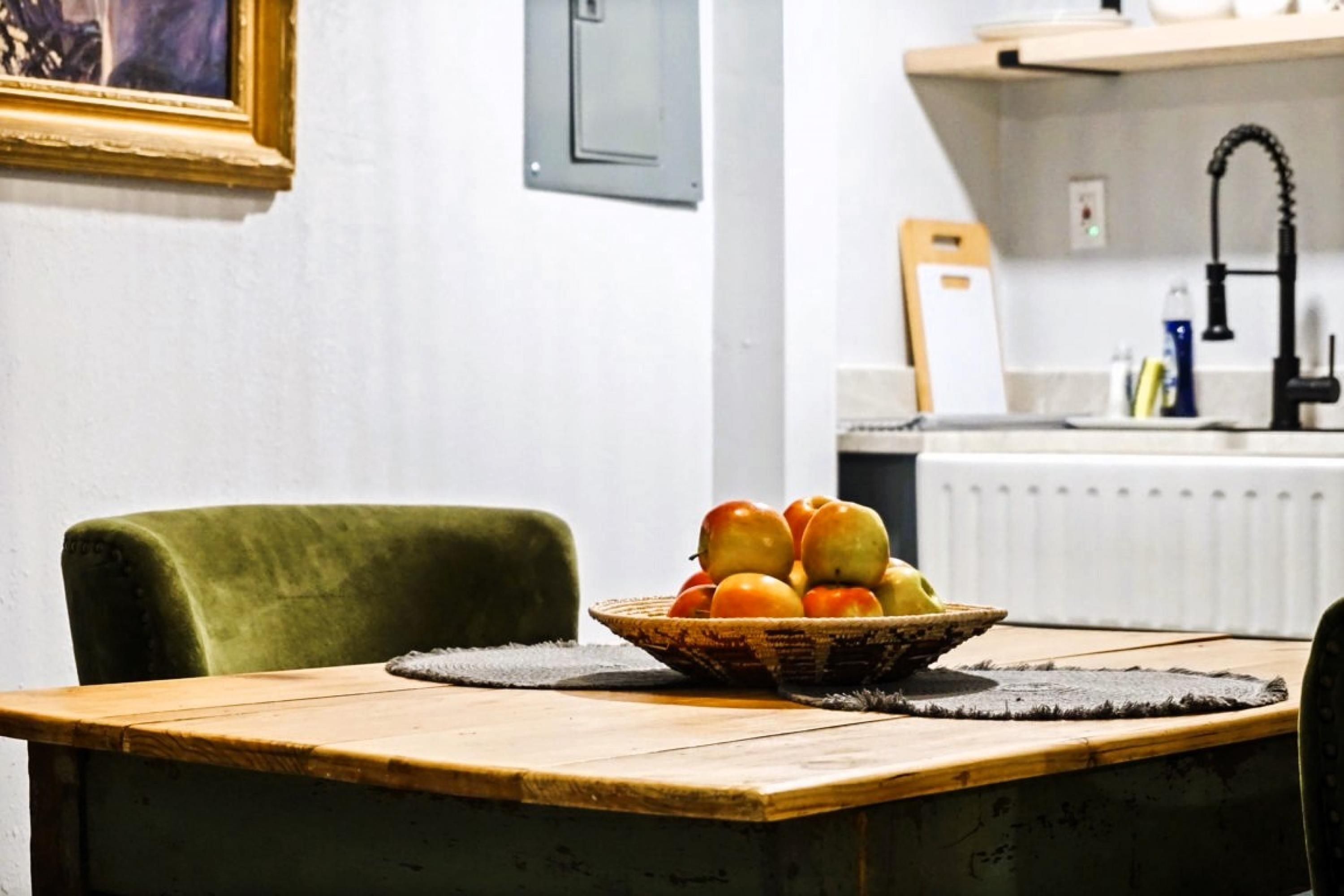 Une table à manger avec un panier de fruits, une chaise verte et un coin cuisine en arrière-plan.