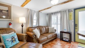 A cozy living room with a brown leather sofa, a blue bee pillow, a small table with a lamp, and a door with a yellow sign. - OIBee Hive • Cute 1970s Beach Cottage 2mi to Beach (Ocean Isle Beach)