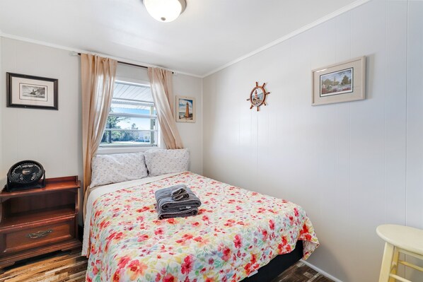 Un dormitorio con colcha estampada, una mesita de noche de madera, un ventilador y una ventana con cortinas.