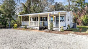 A single-story house with a porch, numbered 6732, surrounded by trees and a gravel driveway. - OIBee Hive • Cute 1970s Beach Cottage 2mi to Beach (Ocean Isle Beach)