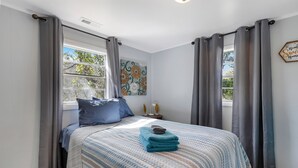 A bedroom with a bed, curtains, a window, a wall art piece, and a bedside table with a lamp. - OIBee Hive • Cute 1970s Beach Cottage 2mi to Beach (Ocean Isle Beach)