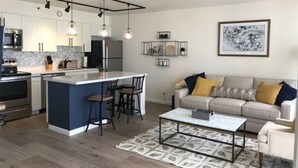 A modern kitchen with a central island, bar stools, and pendant lighting. There is a dining area with a sofa and coffee table.