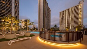 A resort pool area with lounge chairs, a pool, and tall buildings in the background.