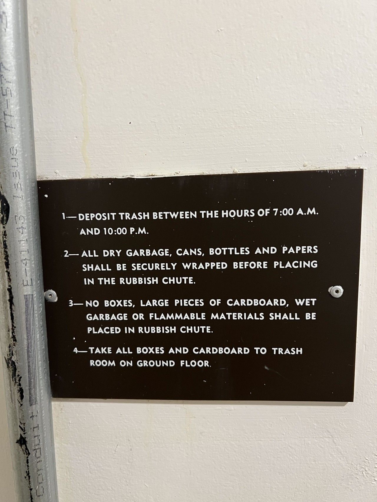 A black sign with white text on a white wall, detailing trash deposit times and guidelines.