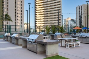 Ulkoilmassa sijaitsevat grillauspaikat, pöydät ja tuolit kaupunkimaisemassa, taustalla korkeita rakennuksia.