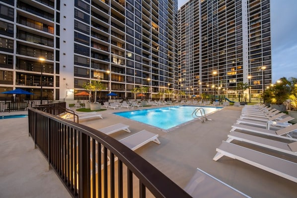 A hotel pool area with sun loungers and a swimming pool, surrounded by high-rise buildings.
