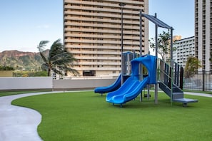 Ein Spielplatz mit zwei Rutschen und einer Kletterkonstruktion, umgeben von grünem Gras und einem Betonweg, mit hohen Gebäuden im Hintergrund.