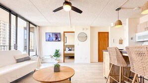 Un salon moderne comprenant un canapé, une table basse ronde, des fauteuils en osier, un ventilateur de plafond, un téléviseur et un coin cuisine.
