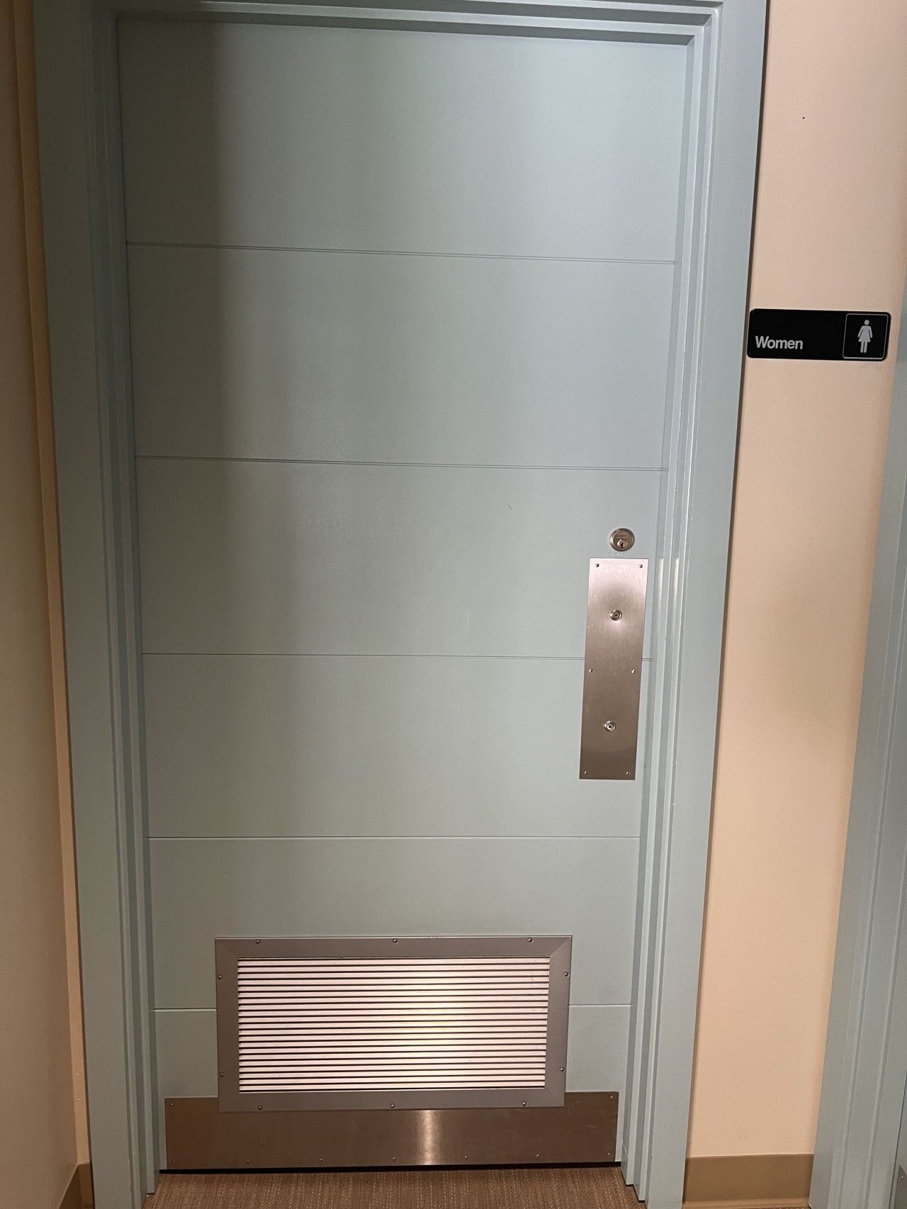 Une porte avec une pancarte indiquant « Femmes », une poignée métallique et une grille d’aération en bas.
