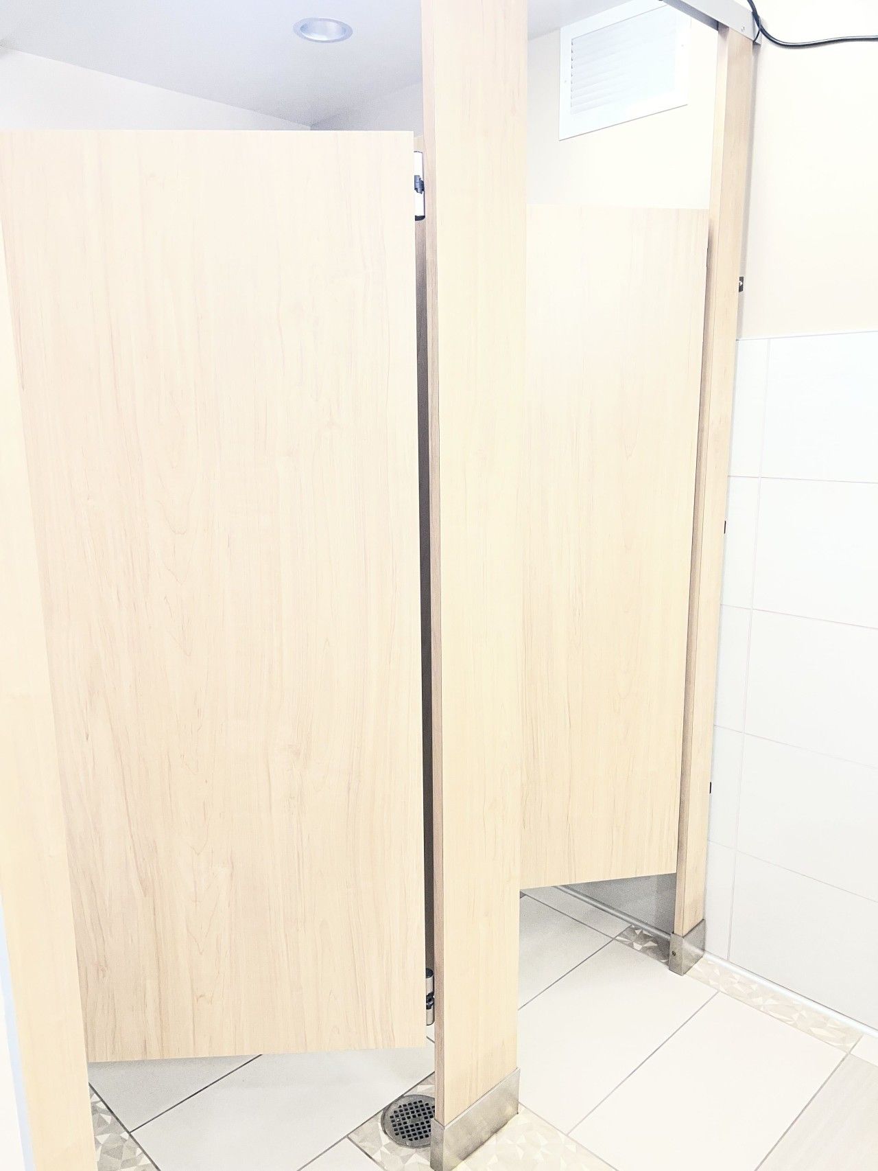 Des toilettes avec des cloisons en bois, un sol carrelé et une grille d’aération visible au plafond.