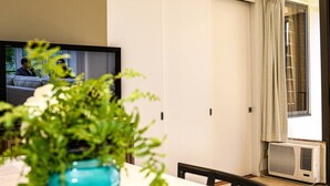 A room with a TV on the wall, a dining table with chairs, a potted plant, and an air conditioning unit.