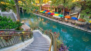 Une promenade au bord de la rivière, avec un escalier en bois descendant vers la rivière, le tout entouré d’une végétation luxuriante et agrémenté de coins salons extérieurs avec des parasols colorés.