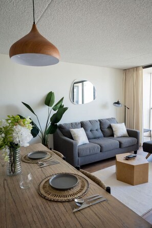 Una sala moderna con un sofá gris, una mesa de centro de madera, una mesa de comedor con platos y cubiertos, una lámpara colgante y un espejo grande en la pared.