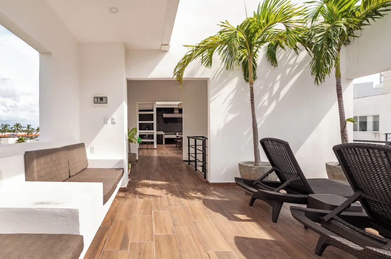Un balcon extérieur moderne avec un plancher en bois, des murs blancs et une vue sur la ville. On y trouve deux fauteuils de jardin en osier et une petite table.