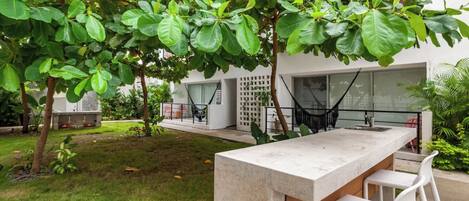 Un área de bar moderna al aire libre con mostrador de concreto y taburetes blancos, rodeada de abundante vegetación y un edificio con amplias ventanas al fondo.