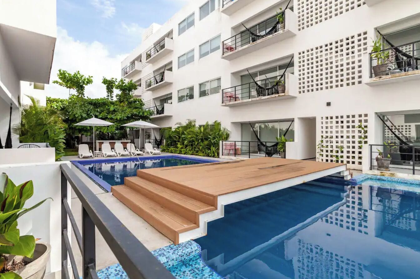 Una piscina en la azotea con escalones de madera, balcones blancos y mobiliario exterior.