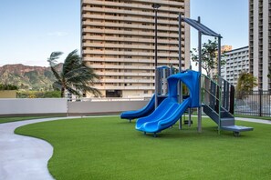 A playground with two slides and a climbing structure, surrounded by green grass and a concrete path, with tall buildings in the background.